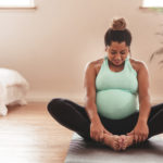 pregnant woman in a mint green tank and black leggings sits on a hardwood floor in butterfly pose