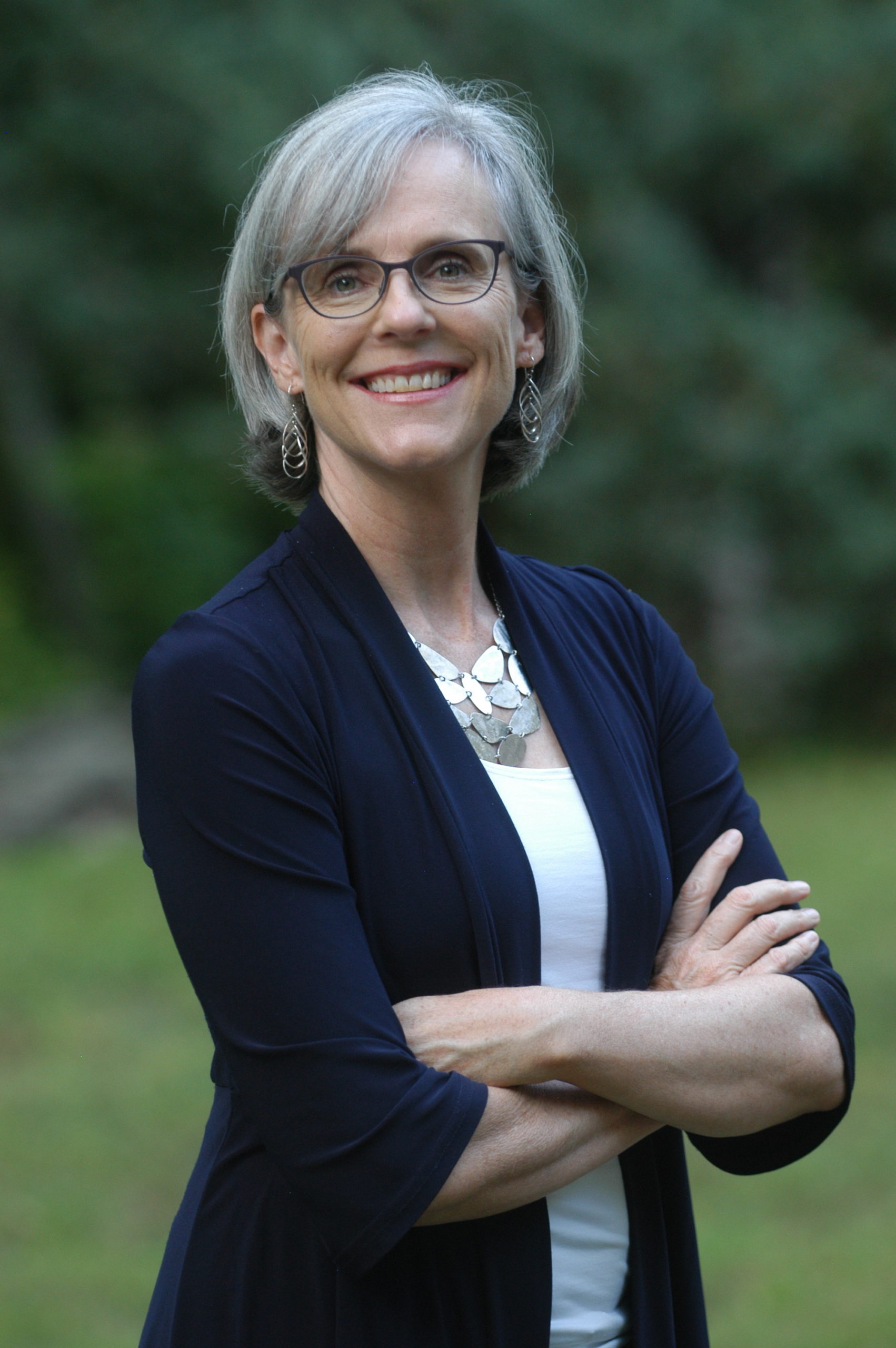 headshot Sage Wheeler, photo of white woman with gray bobbed hair, glasses and wearing silver earring and large silver necklace with navy cardigan and white tank top, standing outdoors
