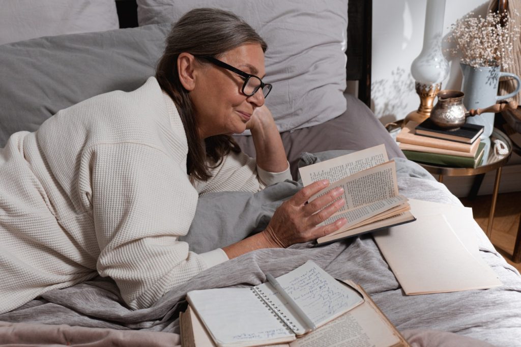 older white woman with thick framed glasses and long gray hair lies on her stomach reading a book in bed