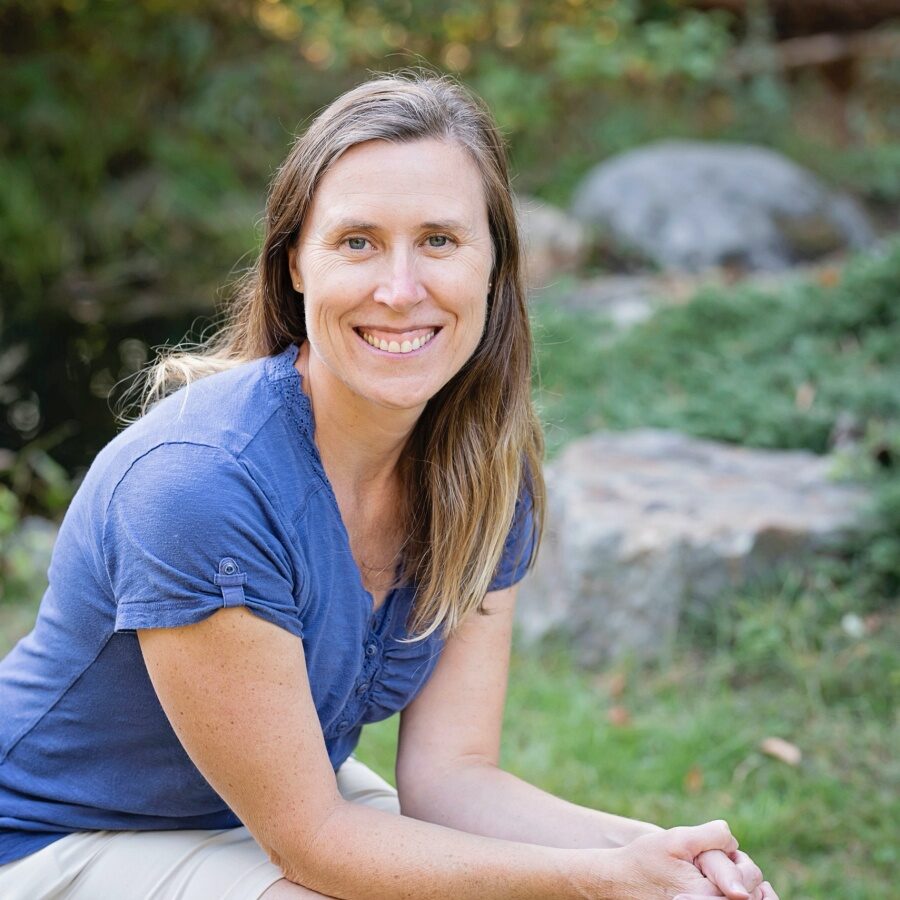 headshot Andrea Mulder, white woman with long blonde hair wearing a blue tee, smiling at the camera, elbows resting on knees in a park