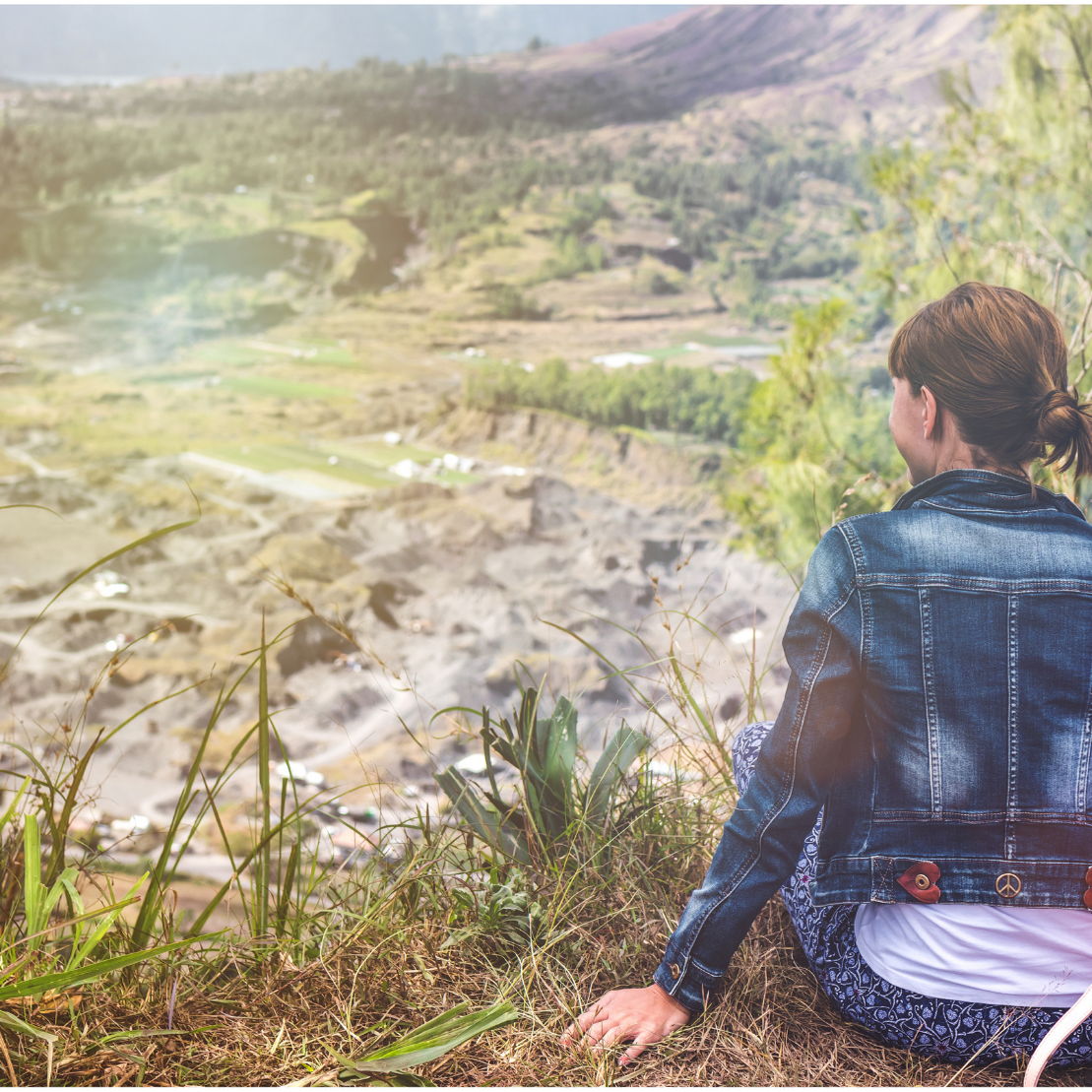 white woman with a messy bun wearing a denim jacket sits looking out over a valley