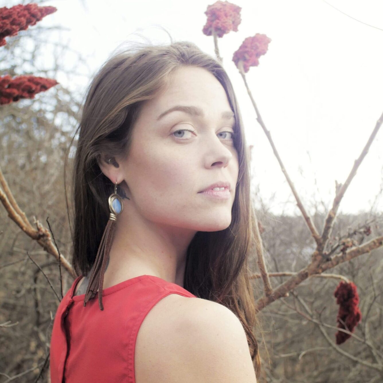 headshot Hope Hartley, woman in red dress with long dark hair looks back at the camera surrounded by bare branches