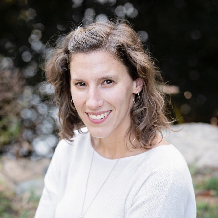 headshot Lisa Colombo, photo of white woman with shoulder-length curly light brown hair, wearing a white tee, sitting in front of a pond