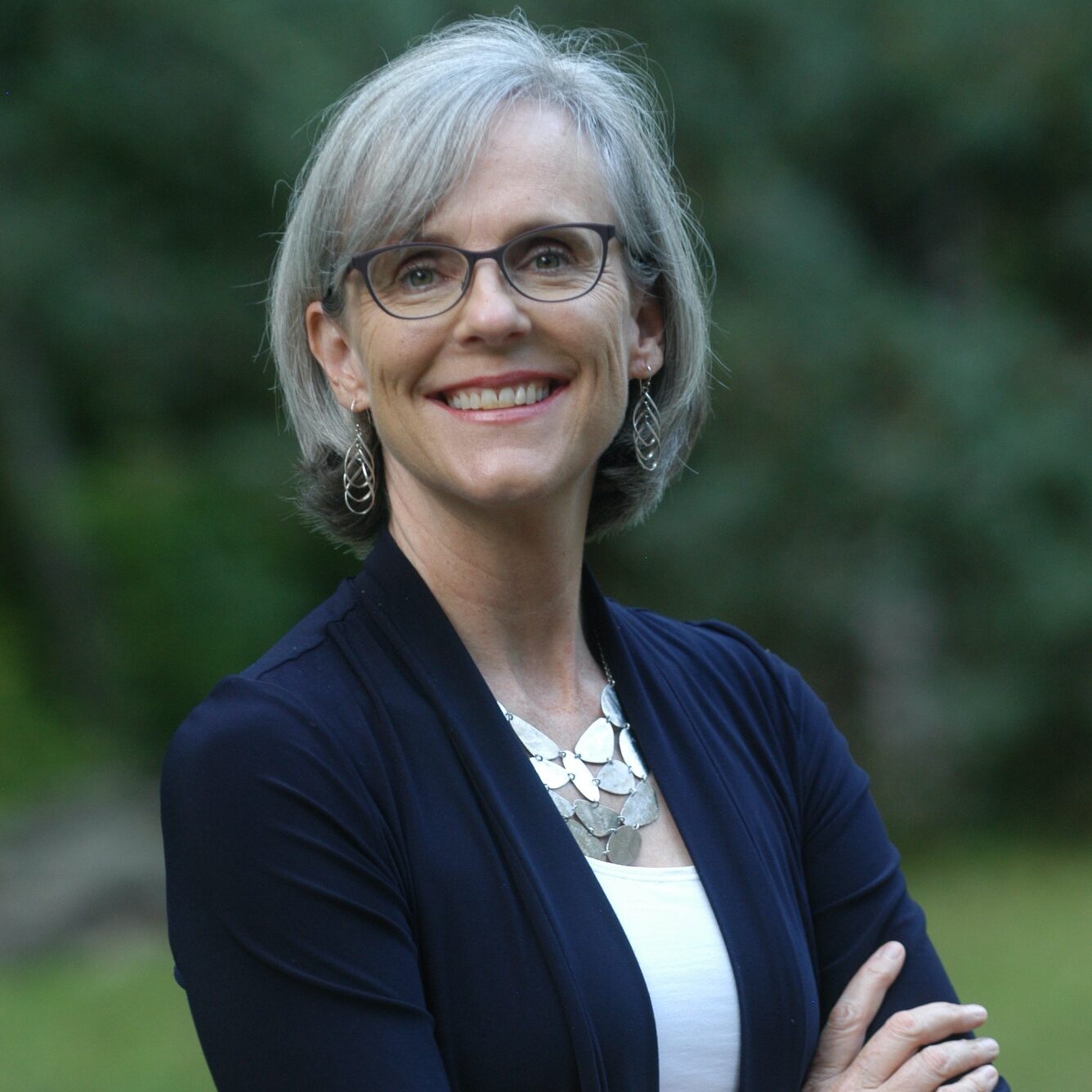 headshot Sage Wheeler, photo of white woman with gray bobbed hair, glasses and wearing silver earring and large silver necklace with navy cardigan and white tank top, standing outdoors