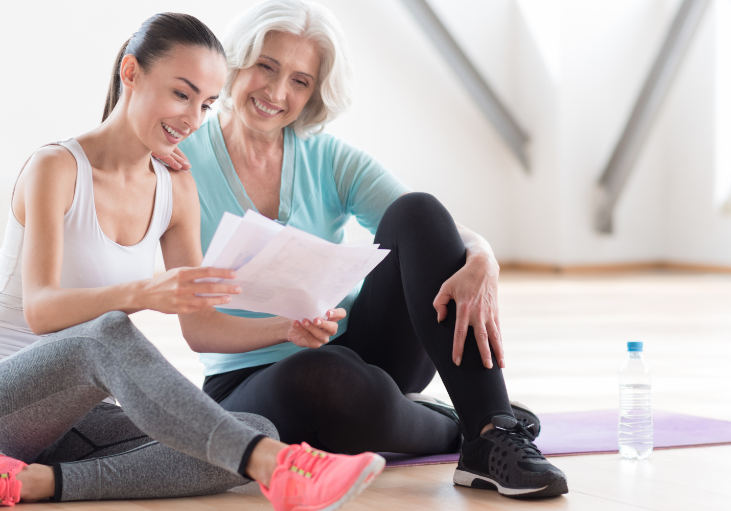 two white women, one older, one younger, sit on a yoga mat reviewing a packet of papers