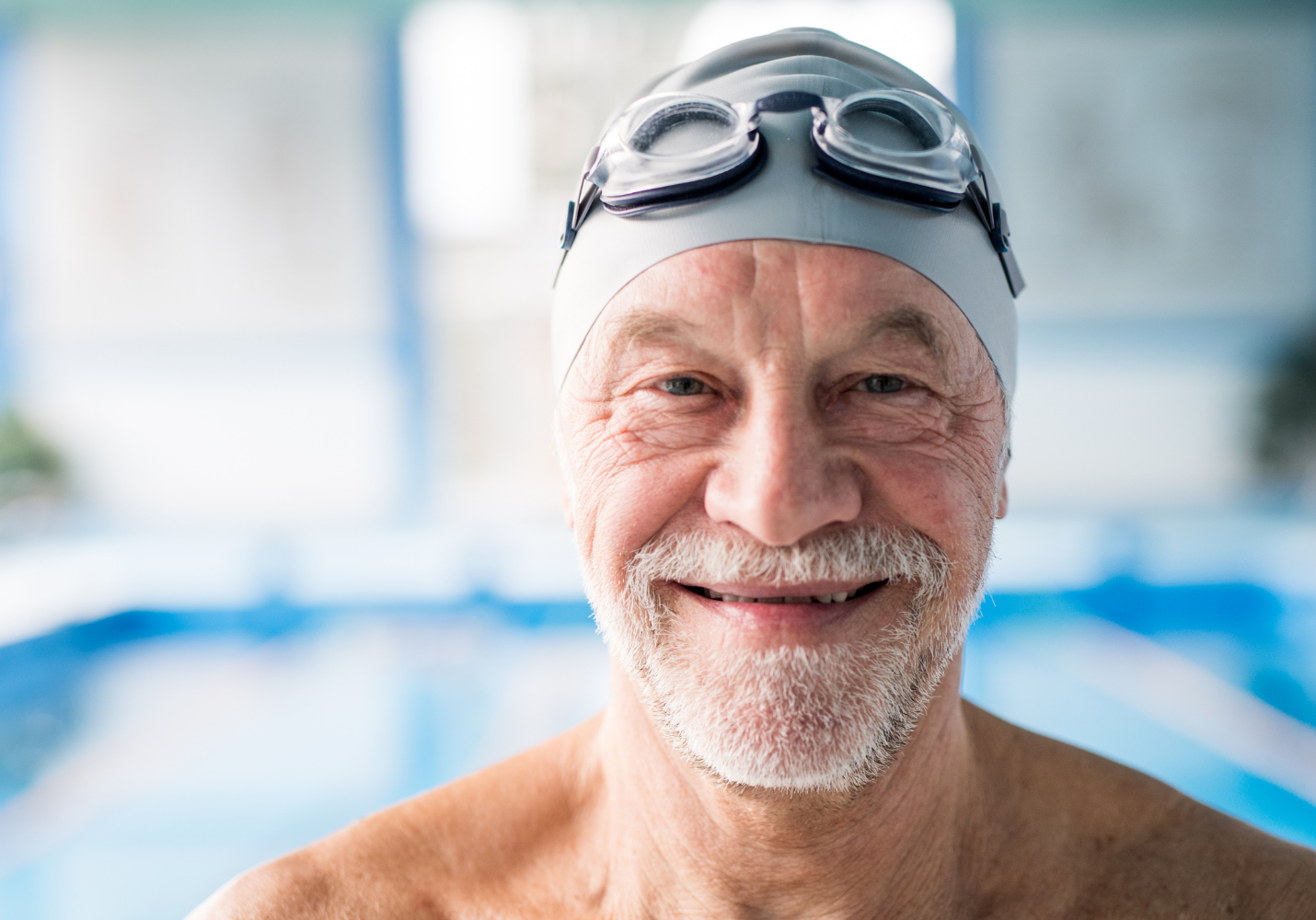close shot of an older white man in swim cap and goggles faces the camera smiling, an indoor pool in the background
