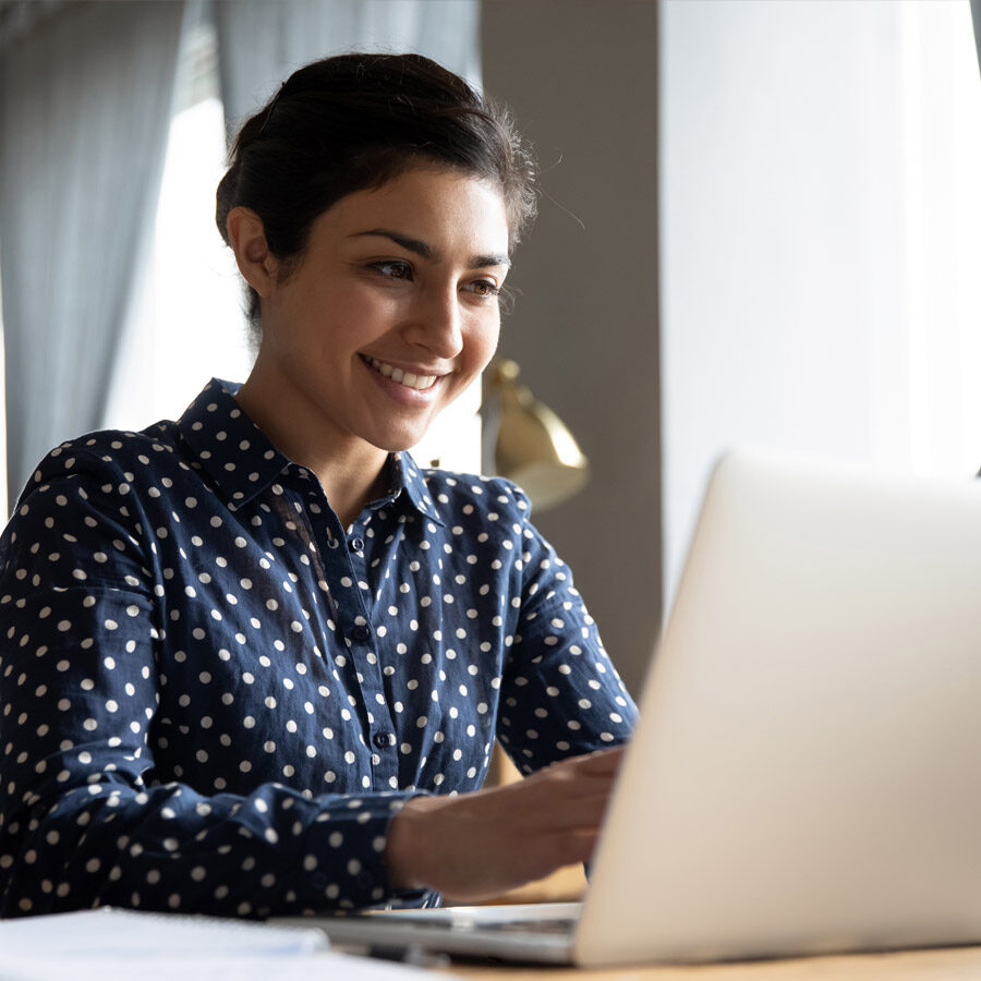 Dark haired woman in a navy polka dot shirt smiles at her laptop