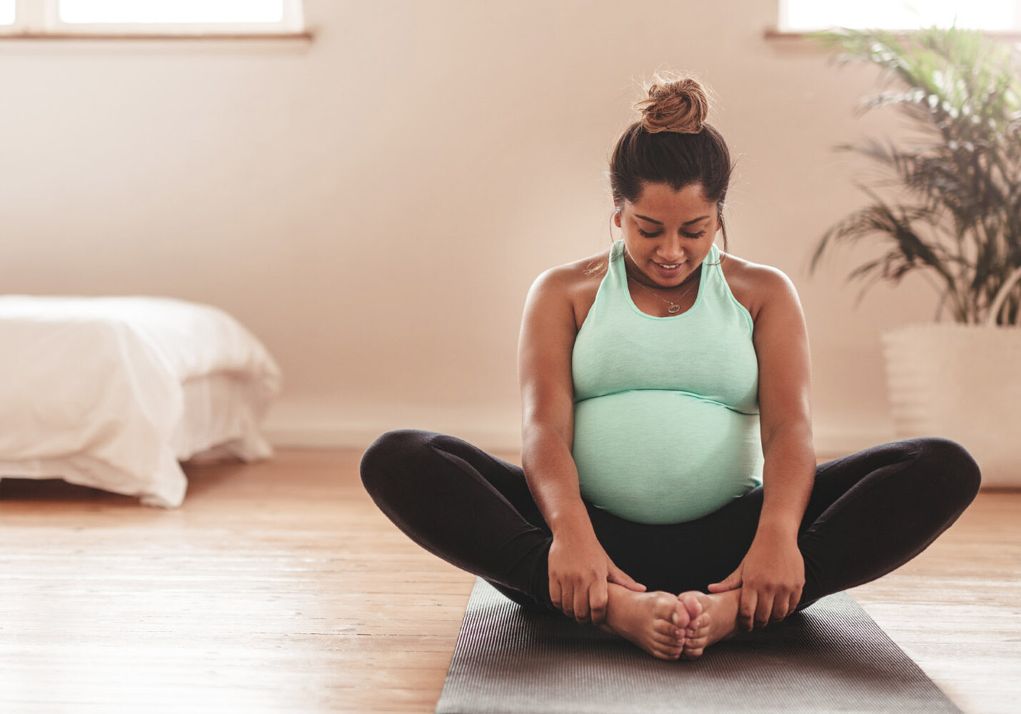 pregnant woman in a mint green tank and black leggings sits on a hardwood floor in butterfly pose