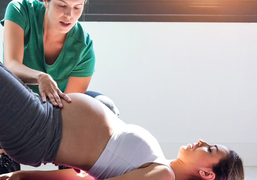pregnant woman in white bra top and leggings lays on her back in bridge pose while another white woman in a green top adjusts her pose with a hand on her belly
